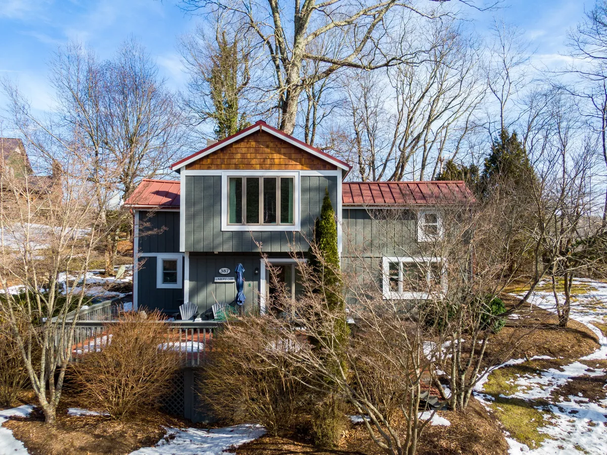 The image shows the charming front facade of a classic Appalachian cabin set amidst tall, tranquil trees. The inviting front porch overlooks the peaceful grove, with cozy seating perfect for relaxing and taking in the serene natural surroundings. Soft sunlight filters through the leaves, creating a warm, welcoming atmosphere that hints at evenings spent watching sunsets or gathering by the fire pit nearby.