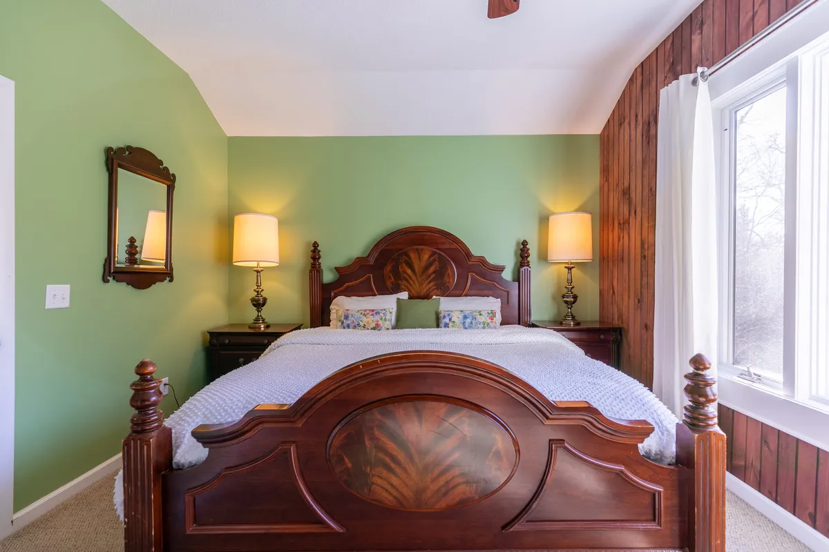 This inviting bedroom features a wooden bed frame with colorful pillows, mirrored wall decor, warm table lamps, and a large window that bathes the room in morning light. The overall mood is warm and welcoming, suitable for a peaceful night's sleep or a quiet read.