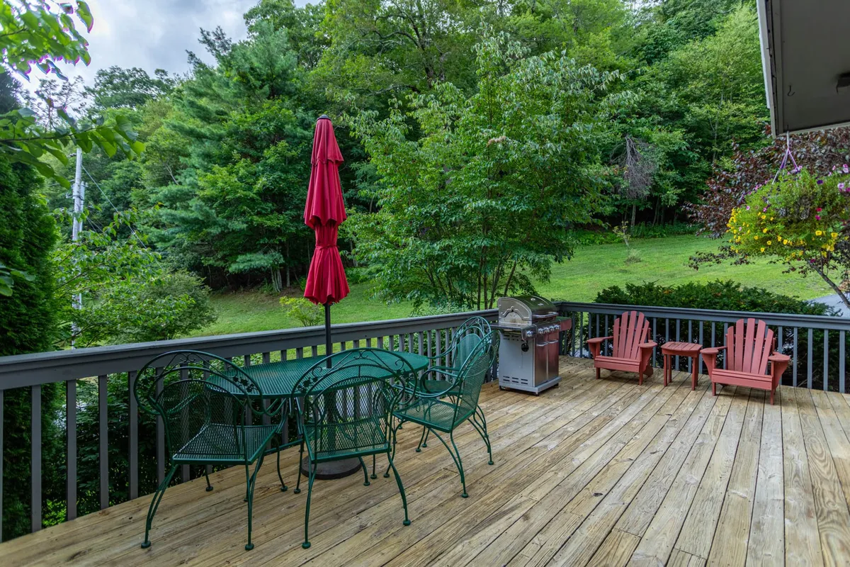 This exterior image features a green dining set with a red umbrella, an outdoor grill, and vibrant red Adirondack chairs, all set on a deck surrounded by lush greenery. The scene is perfect for outdoor dining, barbecuing, and relaxing, with an inviting atmosphere for groups to gather and socialize.