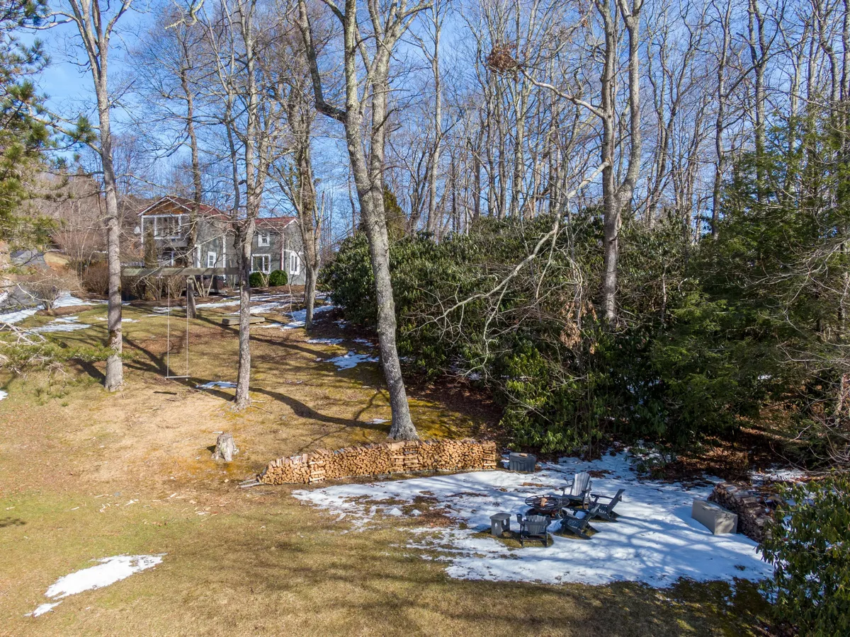 An inviting fire pit area with a generous woodpile is set against a backdrop of a swing set and the house. The scene suggests the warmth of fireside gatherings, opportunities for storytelling, and playful moments in the sloped yard.