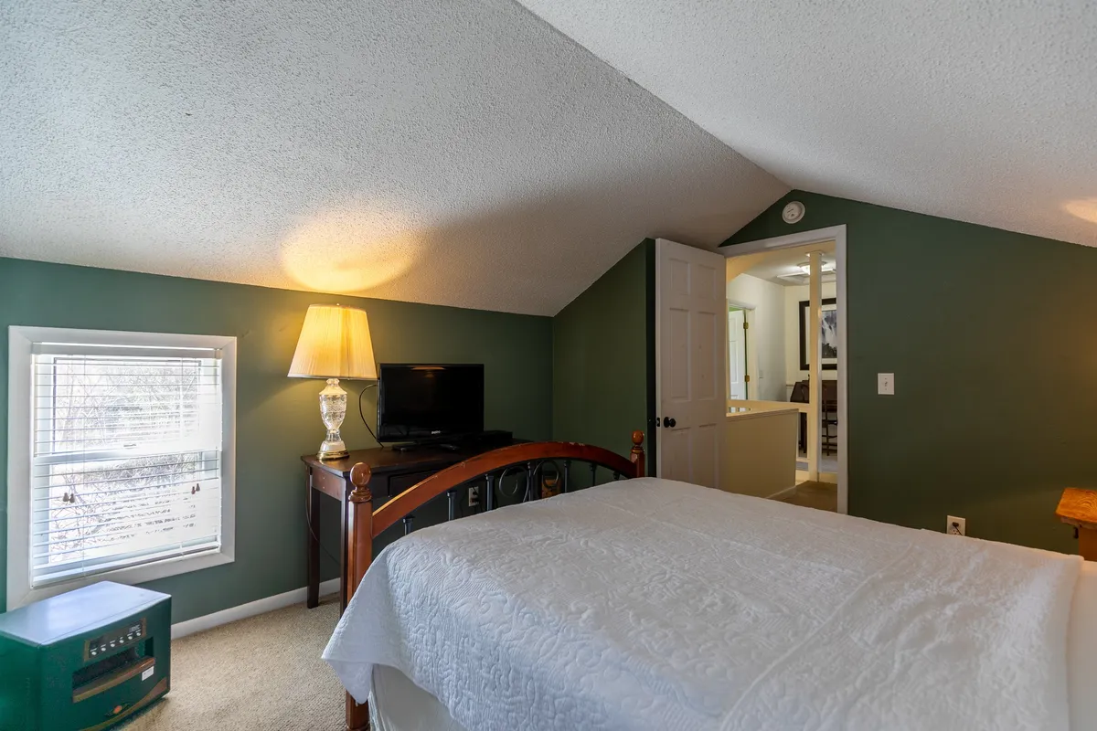 This image presents a peaceful bedroom scene featuring a comfortable bed, a side table with a lamp, a window streaming in natural light, and a television mounted on the wall. The neatly made bed and gentle lighting evoke a sense of serenity and relaxation, ideal for restful nights after a day of adventure.