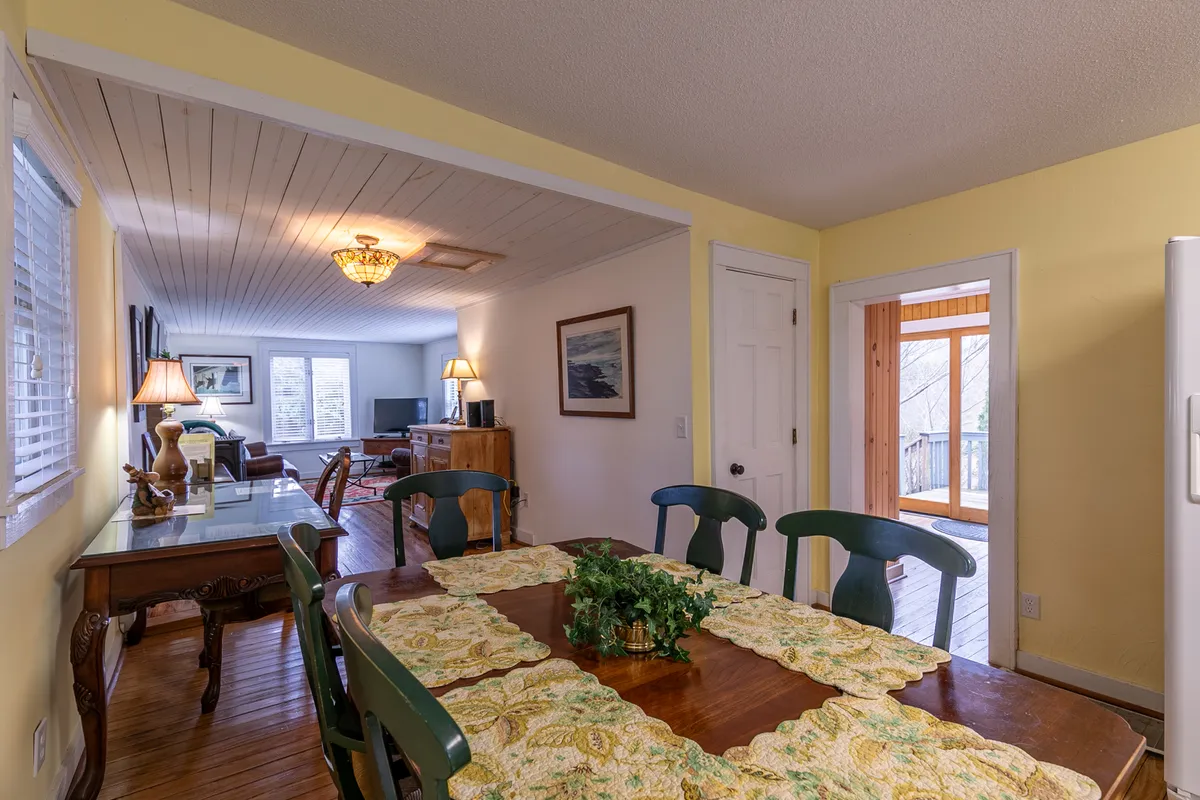 A cozy dining area featuring a table with a decorative runner, softly lit by lamps and natural light streaming through the window. The open doorway connects to the living area, offering a sense of openness and relaxation.
