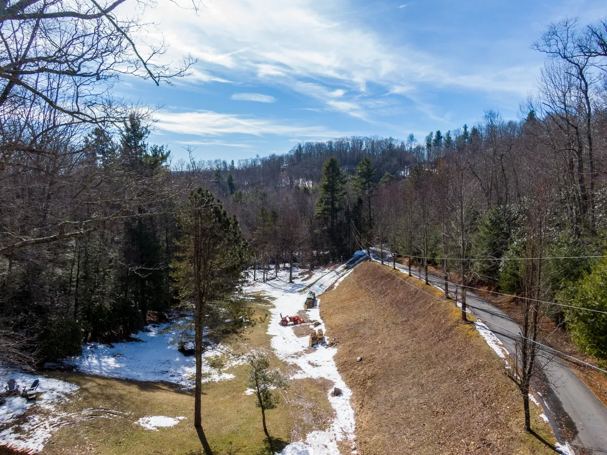 A serene winter landscape showcasing bare trees, patches of lingering snow, a winding road, distant hills, and a hint of construction equipment. The natural setting is quiet and inviting, capturing the transition between seasons and the allure of mountain exploration.