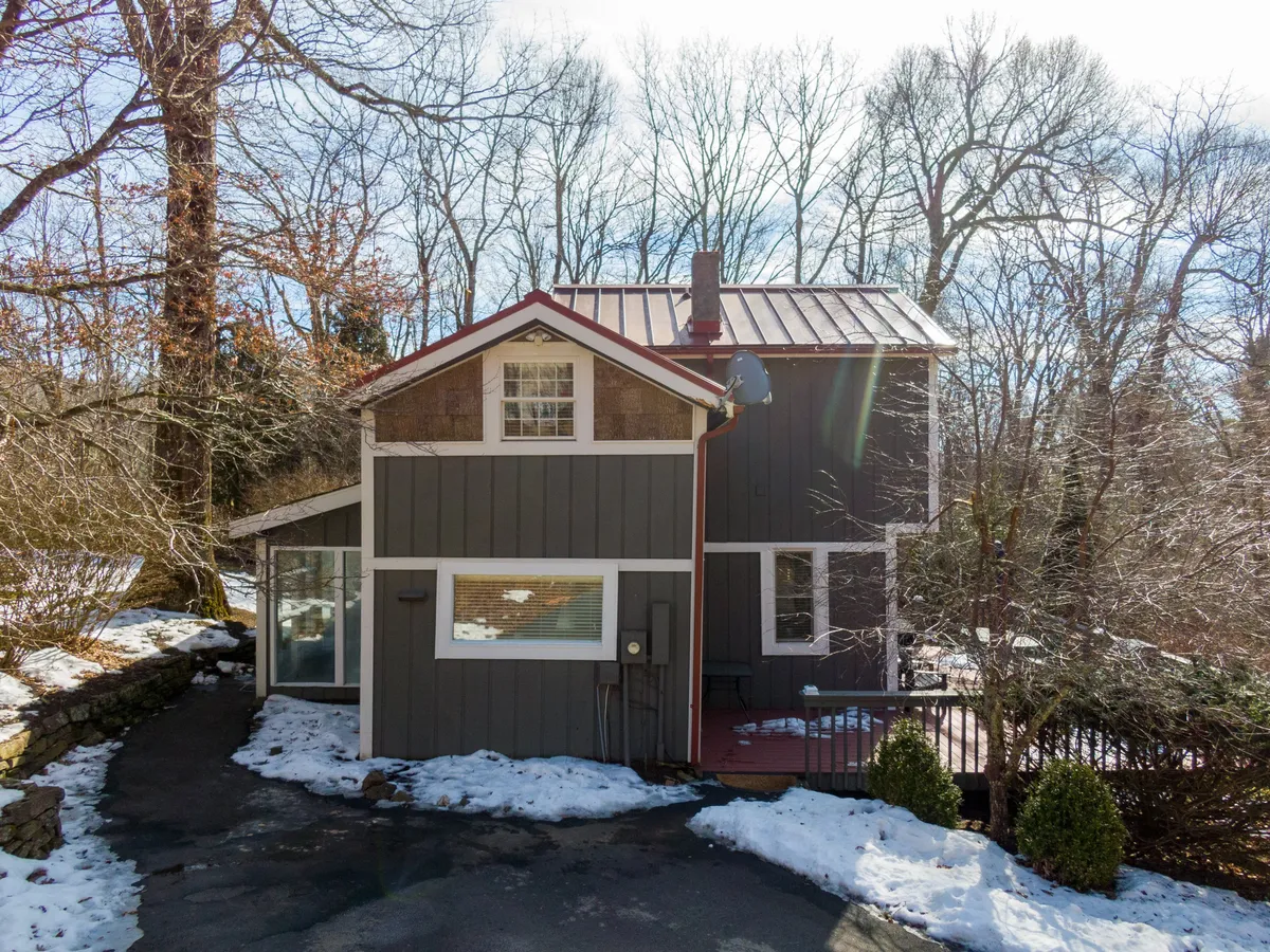 This image captures the exterior of the cabin with its distinctive red metal roof surrounded by lush trees and a driveway leading up to the house. The setting feels inviting and immediately connects the home to the natural landscape that surrounds it.
