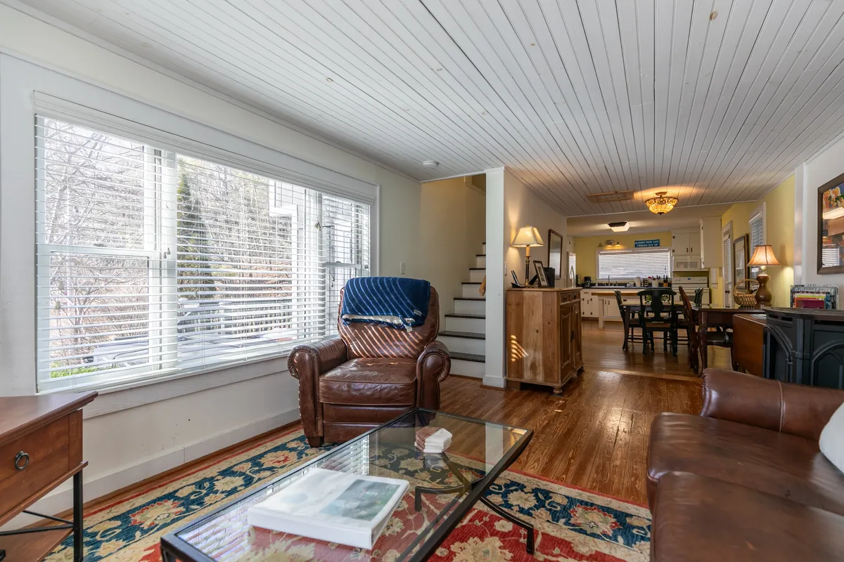 A view that encompasses a plush leather armchair, the dining area with seating for six, a staircase, large window, and a coffee table, all bathed in natural light. The scene feels cozy and ideal for both relaxing and socializing.