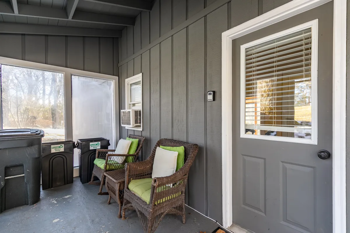 A pair of woven chairs with green cushions sit invitingly on a sunlit porch. Large windows and a door with a glass panel offer views of the outdoors, creating a seamless connection between the inside and nature. The setting suggests a cozy, social atmosphere perfect for sharing drinks or stories after a day of adventure.